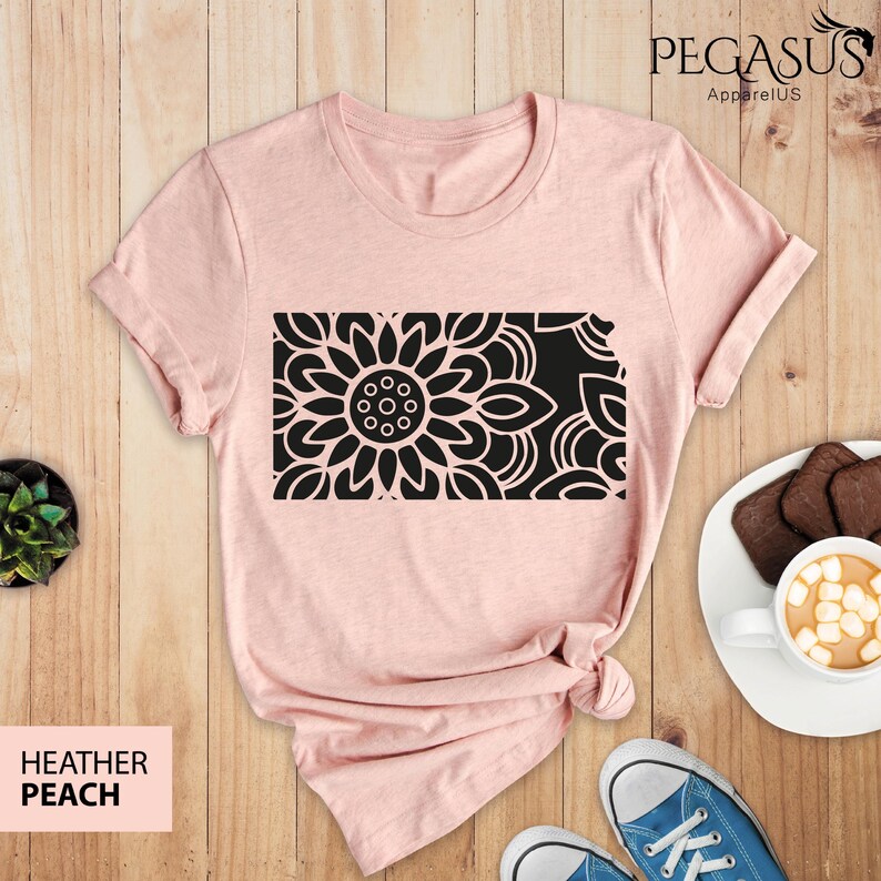May include: A heather peach t-shirt with a black mandala design. The design is a large flower with intricate details. The t-shirt is laid flat on a wooden surface.
