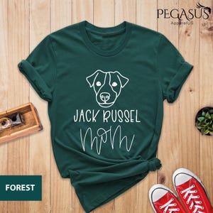 May include: Forest green t-shirt with a white outline of a Jack Russell Terrier's face and the words "JACK RUSSEL MOM" in white script. The shirt is folded and styled on a wooden surface.