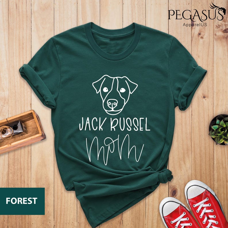 May include: Forest green t-shirt with a white outline of a Jack Russell Terrier's face and the words "JACK RUSSEL MOM" in white script. The shirt is folded and styled on a wooden surface.