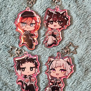 Peut inclure: Quatre porte-clés de personnages d'anime colorés avec un charme en forme d'étoile. Chaque porte-clés présente un personnage différent avec des cheveux noirs, blancs et roux. Les personnages portent des tenues noires et blanches et ont des oreilles de chat.