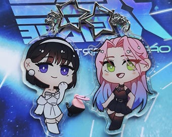 May include: Two anime-style character charms with a silver star-shaped keychain. The charm on the left is a girl with black hair, wearing a white dress and a pink bow. The charm on the right is a girl with pink hair, wearing a black dress and black boots.