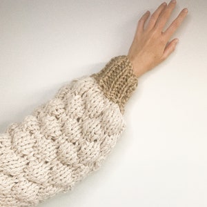 May include: A close-up of a knitted sweater sleeve in beige and brown. The sleeve is made from chunky yarn and has a textured pattern.