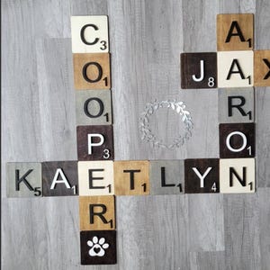 May include: Wooden Scrabble tiles spelling out names: COOPER, JAX, KAETLYN, and JASON. The tiles are various colors, including brown, gray, and cream, with black letters and numbers. A paw print tile is included. A silver wreath is in the center.