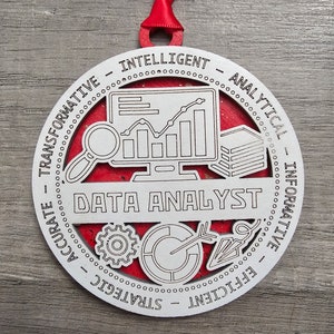 May include: A round white wooden ornament with a red backing and a red ribbon hanger. The center features a computer monitor with a line graph, a magnifying glass, a stack of papers, a gear, a pie chart, and the text DATA ANALYST.