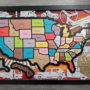 May include: A framed wooden travel map of the United States, featuring individual states in various colors. The map includes cut-out illustrations of transportation modes like a train, airplane, and car. The text "The Wallace's Travel Map" is visible.