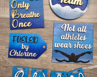 May include: A set of five wooden signs with a blue and white gradient background and gold lettering. The signs feature swim team themed sayings, including "You Only Breathe Once", "Fueled by Chlorine", "Swim Team", "Not all athletes wear shoes", and a banner that reads "Swim".