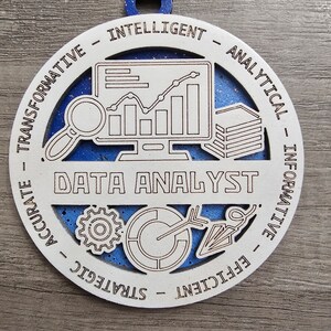 May include: A circular wooden ornament with a blue glitter background and white laser-cut overlay. The center features the text DATA ANALYST with icons of a computer monitor, magnifying glass, gear, and chart. The border includes the words TRANSFORMATIVE, INTELLIGENT, ANALYTICAL, INFORMATIVE, EFFICIENT, STRATEGIC, and ACCURATE.