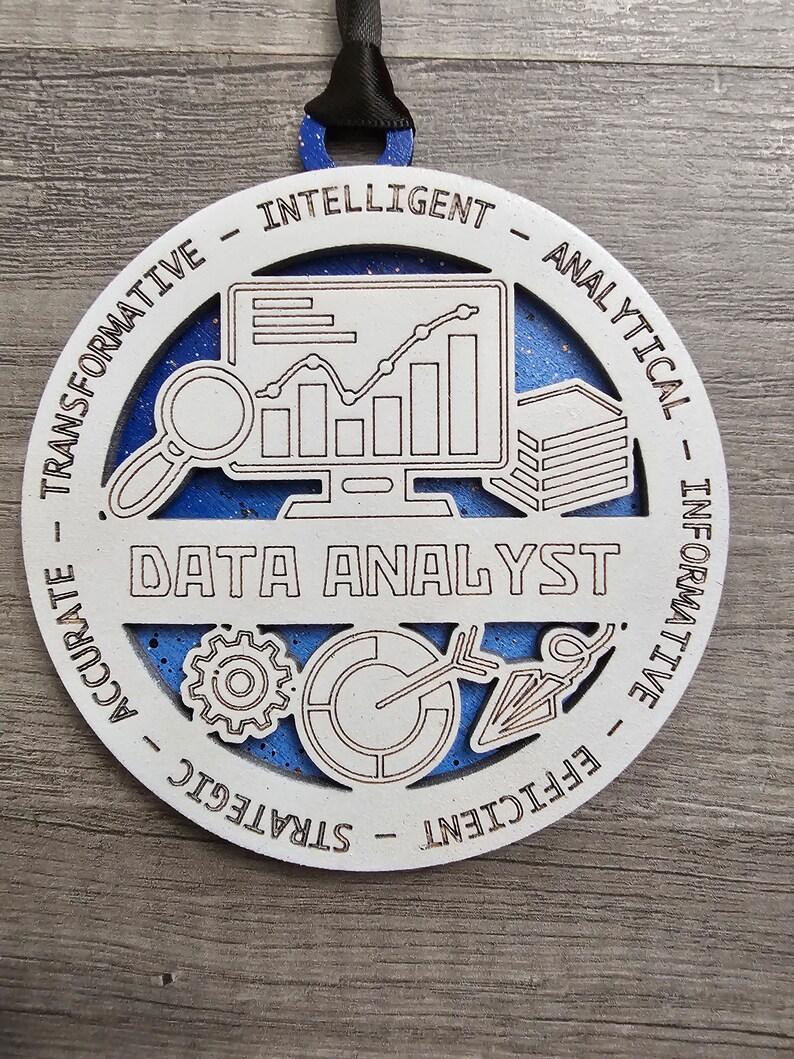 May include: A circular wooden ornament with a blue glitter background and white laser-cut overlay. The center features the text DATA ANALYST with icons of a computer monitor, magnifying glass, gear, and chart. The border includes the words TRANSFORMATIVE, INTELLIGENT, ANALYTICAL, INFORMATIVE, EFFICIENT, STRATEGIC, and ACCURATE.