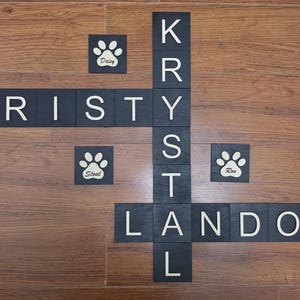 May include: A set of black square wooden tiles with white letters arranged in a crossword pattern. The tiles spell out the names KRISTY, KRYSTAL, and LANDON. Three additional tiles feature white paw prints with the names Daisy, Stout, and Roo.