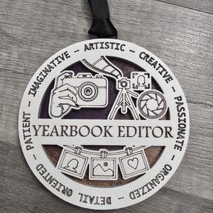 May include: White wooden ornament with a black ribbon. The ornament features a camera, tripod, and lens with the words "Yearbook Editor" in the center. The words "Patient", "Imaginative", "Artistic", "Creative", "Passionate", "Organized", "Detail Oriented" are written around the edge of the ornament.