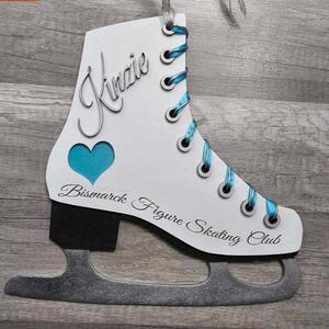 May include: A white ice skate ornament with teal blue laces and a teal heart cutout. The name "Kinzie" is scripted on the side, and the text "Bismarck Figure Skating Club" is printed below.