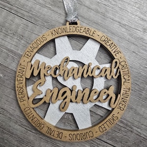 May include: A wooden ornament with the words "Mechanical Engineer" in a cursive font, set within a gear-shaped design. The ornament includes the words "Tenacious," "Knowledgeable," "Creative," and other traits. A silver ribbon is attached for hanging.