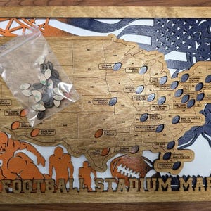 May include: A wooden map of the United States featuring laser-cut football stadium names, an orange and navy blue layered background with football player silhouettes, and a clear plastic bag containing small wooden football-shaped markers.