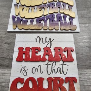 May include: Two wooden signs with a volleyball theme. The top sign has the word "VOLLEYBALL" written three times in gold and purple letters. The bottom sign has the words "my heart is on that court" written in red and gold letters.