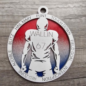 May include: A white wooden ornament shaped like a football player with the name "Wallin" and the number "67" on the back. The ornament is decorated with a red, white, and blue gradient and features the words "Hail Mary", "Punt", "Touch Down", "Field Goal", "Interception", "Red Zone", and "Audible Blitz" around the edge.