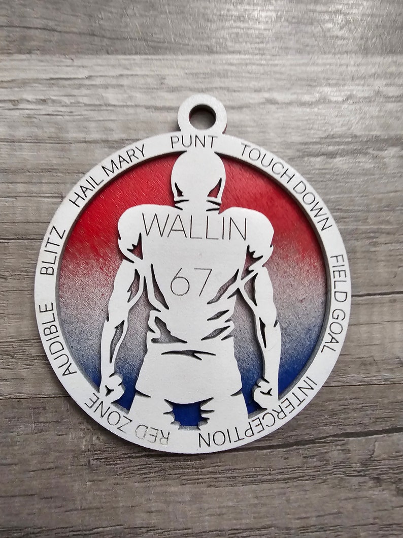 May include: A white wooden ornament shaped like a football player with the name "Wallin" and the number "67" on the back. The ornament is decorated with a red, white, and blue gradient and features the words "Hail Mary", "Punt", "Touch Down", "Field Goal", "Interception", "Red Zone", and "Audible Blitz" around the edge.