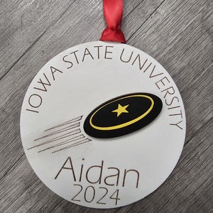 May include: A white wooden ornament with the text "IOWA STATE UNIVERSITY" and "Aidan 2024". The ornament features a black and yellow design of a flying object with a star. A red ribbon is attached for hanging.