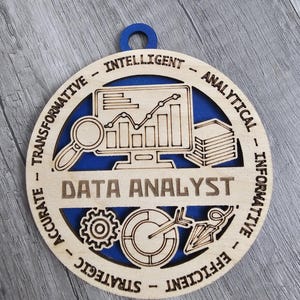 May include: A circular wooden ornament with a blue backing, featuring laser-cut designs of a computer monitor with a line graph, a magnifying glass, a stack of papers, a gear, a pie chart, and an arrow. The text reads DATA ANALYST, INTELLIGENT, ANALYTICAL, INFORMATIVE, EFFICIENT, STRATEGIC, ACCURATE, and TRANSFORMATIVE.