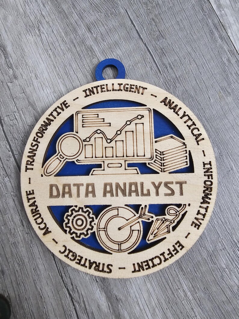 May include: A circular wooden ornament with a blue backing, featuring laser-cut designs of a computer monitor with a line graph, a magnifying glass, a stack of papers, a gear, a pie chart, and an arrow. The text reads DATA ANALYST, INTELLIGENT, ANALYTICAL, INFORMATIVE, EFFICIENT, STRATEGIC, ACCURATE, and TRANSFORMATIVE.