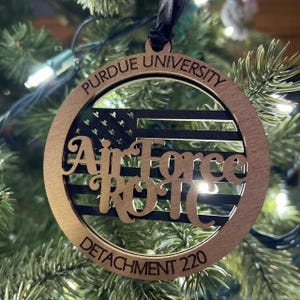 May include: A circular wooden ornament featuring a black flag design with stars and stripes. The text PURDUE UNIVERSITY is engraved at the top, Air Force ROTC in the center, and DETACHMENT 220 at the bottom. It hangs from a black ribbon.