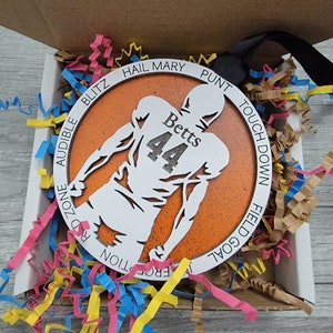 May include: A round orange and white wooden ornament featuring a silhouette of a football player. The ornament is inscribed with the words "Hail Mary", "Punt", "Touch Down", "Field Goal", "Red Zone", "Blitz", "Audible", "Interception", and "Fumble". The player has the name "Betts" and the number "44" on the back of their jersey.
