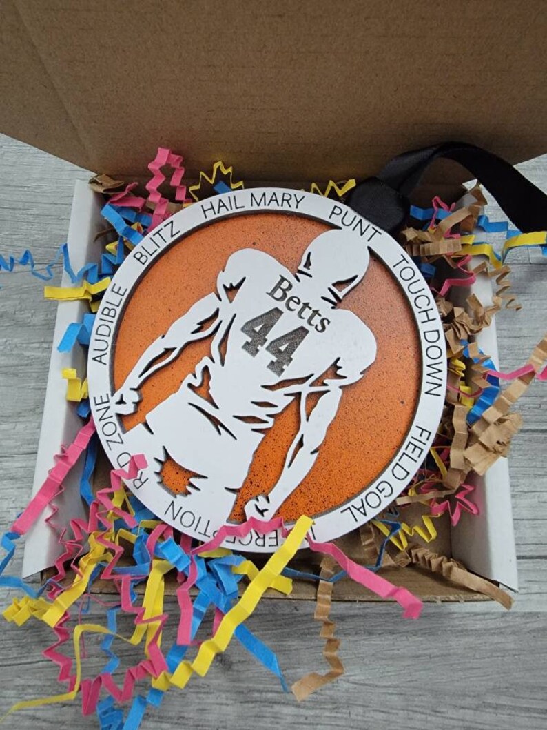 May include: A round orange and white wooden ornament featuring a silhouette of a football player. The ornament is inscribed with the words "Hail Mary", "Punt", "Touch Down", "Field Goal", "Red Zone", "Blitz", "Audible", "Interception", and "Fumble". The player has the name "Betts" and the number "44" on the back of their jersey.