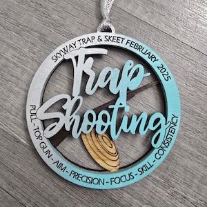 May include: A wooden Trap Shooting ornament with a silver ribbon for hanging. The ornament features the text "Trap Shooting" in a stylized font, with additional text around the edge, including "Skyway Trap & Skeet February 2025".
