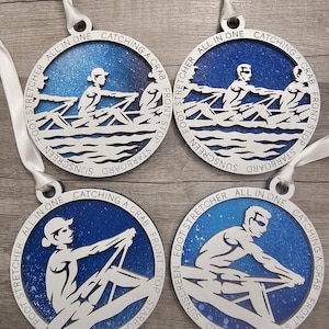 May include: Four round, white wooden ornaments with blue and white designs. Each ornament features a silhouette of a person rowing a boat with the text "ALL IN ONE CATCHING A CRAB FRONT STOP STARBOARD SUNSCREEN FOOT STRETCHER" around the edge.