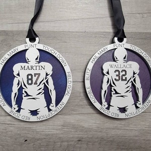 May include: Two white wooden football player ornaments with purple backgrounds. The ornaments feature the names "Martin" and "Wallace" with the numbers "87" and "32" on the back of the players. The ornaments have a variety of football terms around the edge.