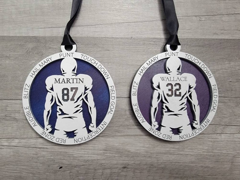 May include: Two white wooden football player ornaments with purple backgrounds. The ornaments feature the names "Martin" and "Wallace" with the numbers "87" and "32" on the back of the players. The ornaments have a variety of football terms around the edge.