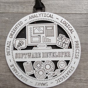 May include: A white, circular wooden ornament with the words "SOFTWARE DEVELOPER" in the center. The ornament features cut-out graphics of a computer, gears, and other tech-related icons. The words "ANALYTICAL", "LOGICAL", "PRECISE", "STRATEGIC", "SMART", and "ADAPTABLE" are around the edge.