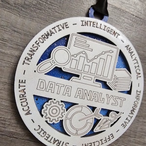 May include: A circular white wooden ornament with a blue glittery background, featuring the text DATA ANALYST in the center. Icons include a computer monitor with a line graph, a magnifying glass, a gear, a pie chart, and an arrow. The border displays the words TRANSFORMATIVE, INTELLIGENT, ANALYTICAL, INFORMATIVE, EFFICIENT, STRATEGIC, and ACCURATE.