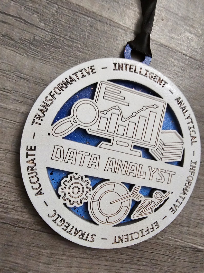 May include: A circular white wooden ornament with a blue glittery background, featuring the text DATA ANALYST in the center. Icons include a computer monitor with a line graph, a magnifying glass, a gear, a pie chart, and an arrow. The border displays the words TRANSFORMATIVE, INTELLIGENT, ANALYTICAL, INFORMATIVE, EFFICIENT, STRATEGIC, and ACCURATE.