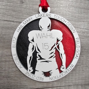 May include: A white and red round wooden ornament with a silhouette of a football player. The ornament has the words "Hail Mary Lakers Touchdown", "Audible Blitz", "Red Zone", "Interception", "Field Goal" and "Wahl 15" printed around the edge.
