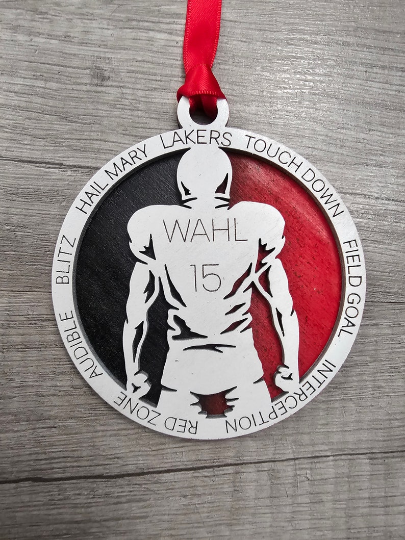 May include: A white and red round wooden ornament with a silhouette of a football player. The ornament has the words "Hail Mary Lakers Touchdown", "Audible Blitz", "Red Zone", "Interception", "Field Goal" and "Wahl 15" printed around the edge.
