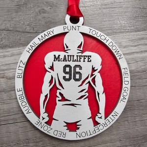May include: A white wooden ornament shaped like a football player with the name "M.CAULIFFE" and the number "96" on the back. The ornament is red and has the words "HAIL MARY", "PUNT", "TOUCHDOWN", "FIELD GOAL", "INTERCEPTION", "RED ZONE", "AUDIBLE", and "BLITZ" around the edge.