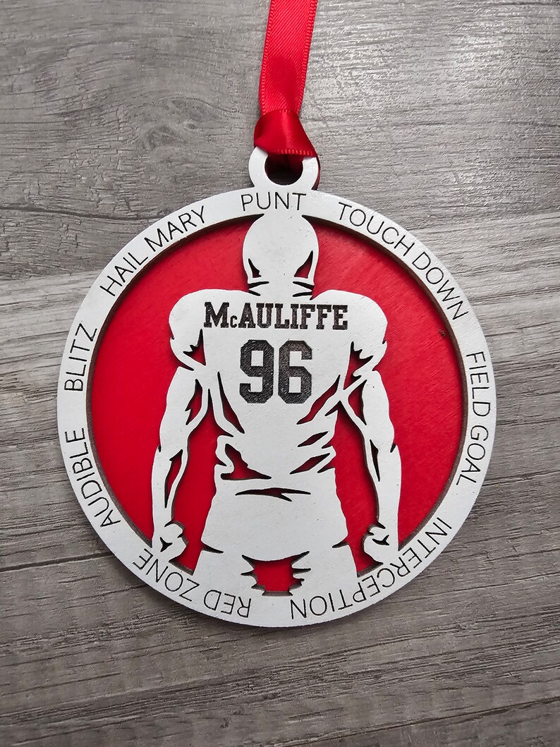 May include: A white wooden ornament shaped like a football player with the name "M.CAULIFFE" and the number "96" on the back. The ornament is red and has the words "HAIL MARY", "PUNT", "TOUCHDOWN", "FIELD GOAL", "INTERCEPTION", "RED ZONE", "AUDIBLE", and "BLITZ" around the edge.