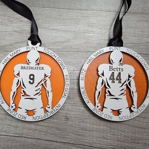 May include: Two orange and white wooden football ornaments with the names "Betts" and "Breimayer" and their corresponding jersey numbers, 44 and 9, printed on them. The ornaments are decorated with football terms around the edges.