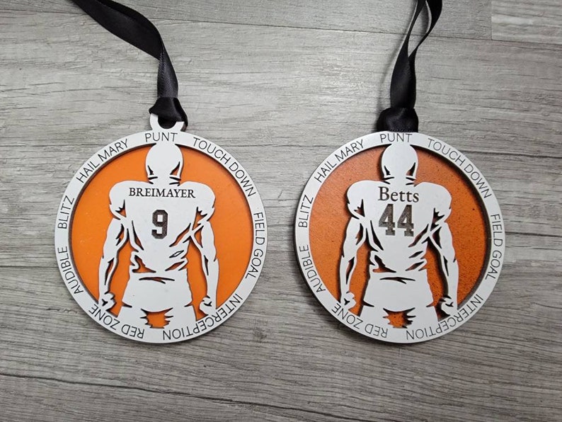 May include: Two orange and white wooden football ornaments with the names "Betts" and "Breimayer" and their corresponding jersey numbers, 44 and 9, printed on them. The ornaments are decorated with football terms around the edges.