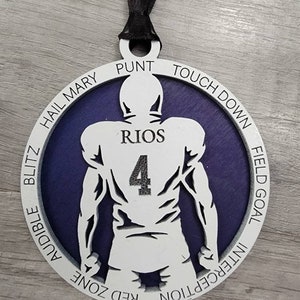 May include: A white wooden football player ornament with a purple background. The player is wearing a jersey with the number 4 and the name "RIOS" on the back. The ornament has a black ribbon for hanging and the words "TOUCHDOWN", "FIELD GOAL", "INTERCEPTION", "RED ZONE", "AUDIBLE", "BLITZ", "HAIL MARY", and "PUNT" around the edge.