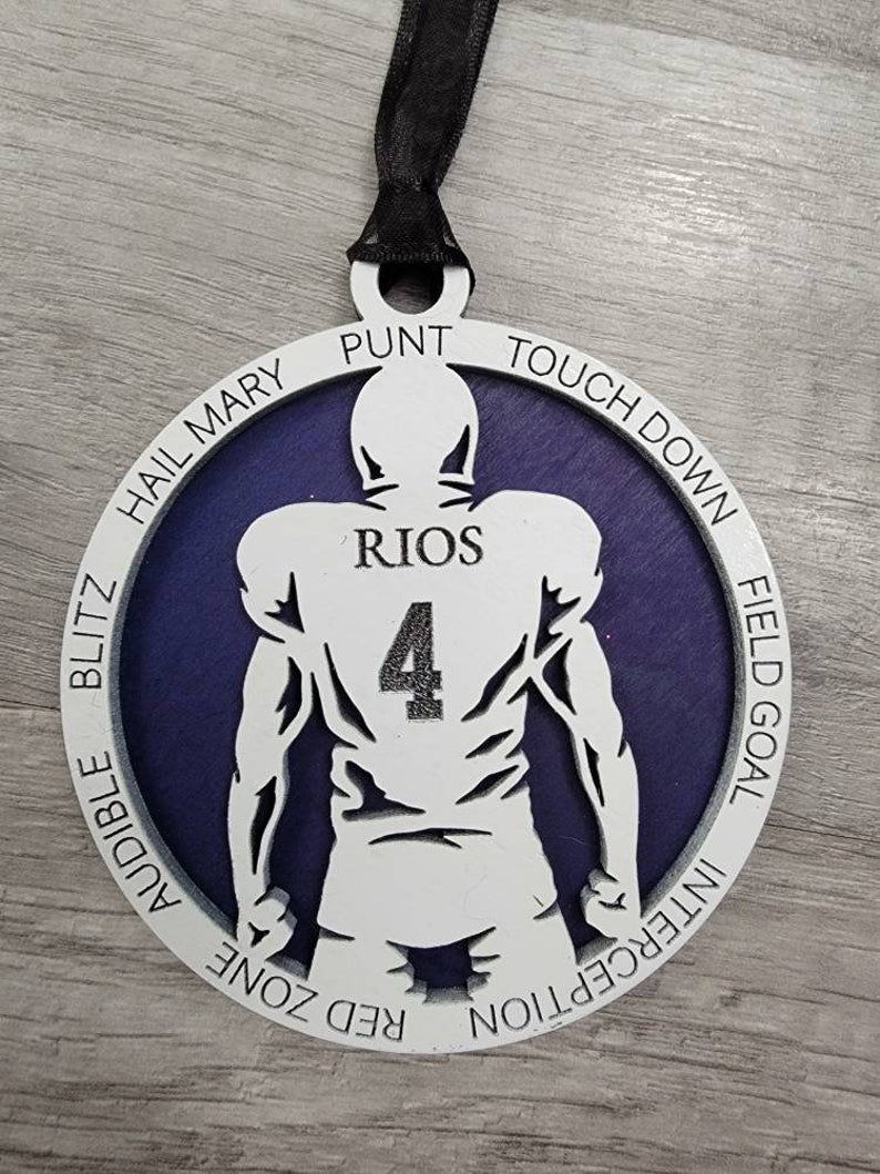 May include: A white wooden football player ornament with a purple background. The player is wearing a jersey with the number 4 and the name "RIOS" on the back. The ornament has a black ribbon for hanging and the words "TOUCHDOWN", "FIELD GOAL", "INTERCEPTION", "RED ZONE", "AUDIBLE", "BLITZ", "HAIL MARY", and "PUNT" around the edge.