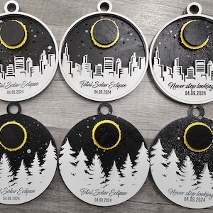 May include: Six round, white, wooden ornaments with black and yellow designs. The ornaments feature a black silhouette of a city skyline with a yellow crescent moon and the text "Total Solar Eclipse 04.08.2024" or "Never stop looking up 04.08.2024".