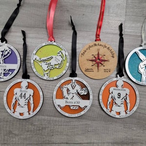 May include: Seven round wooden decorations with various sports themes. The decorations are painted in different colours, including orange, blue, green, and brown. Each decoration features a different sport, including football, gymnastics, swimming, and hockey. The decorations are decorated with text and numbers, such as "Betts #30", "Gomes", and "Breimayer 9".