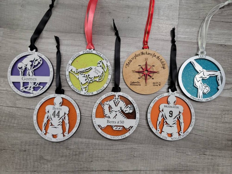 May include: Seven round wooden decorations with various sports themes. The decorations are painted in different colours, including orange, blue, green, and brown. Each decoration features a different sport, including football, gymnastics, swimming, and hockey. The decorations are decorated with text and numbers, such as "Betts #30", "Gomes", and "Breimayer 9".