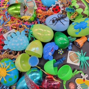May include: A collection of colorful plastic Easter eggs filled with small toys, including a blue ant, a green frog, a yellow spider, a red ladybug, a blue car, and a green and yellow bee. The eggs are surrounded by shredded paper.