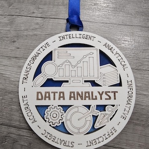 May include: A white wooden ornament with a blue background and the words "Data Analyst" in the center. The ornament is decorated with icons representing data analysis, such as a magnifying glass, a computer screen with a graph, and a stack of books. The words "Transformative", "Intelligent", "Analytical", "Informative", "Accurate", "Efficient", "Strategic" are written around the edge of the ornament.