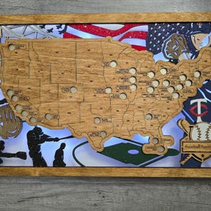 May include: A rectangular wooden wall map of the United States featuring laser-cut state borders and circular cutouts for baseball stadiums. The design includes a baseball player illustration, a baseball graphic, and the text Baseball Travel Map.