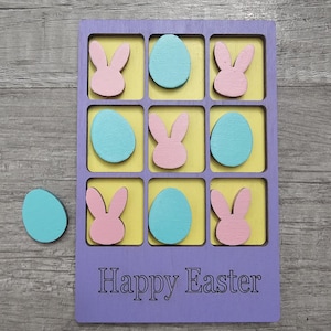 May include: A wooden Easter-themed tic-tac-toe game. The game board is purple with yellow squares, featuring pink bunny and blue egg game pieces. The words "Happy Easter" are engraved on the board. An extra blue egg is next to the board.
