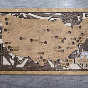 May include: Wooden soccer travel map of the United States, laser-cut with stadium locations. The map is framed in a light brown wood, with a darker wood border and accents. The text "SOCCER TRAVEL MAP" is visible.
