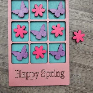 May include: A pink wooden game board with a grid of nine squares, each containing a teal background. The squares feature purple butterflies and pink flowers. The words "Happy Spring" are printed at the bottom. A loose pink flower is next to the board.